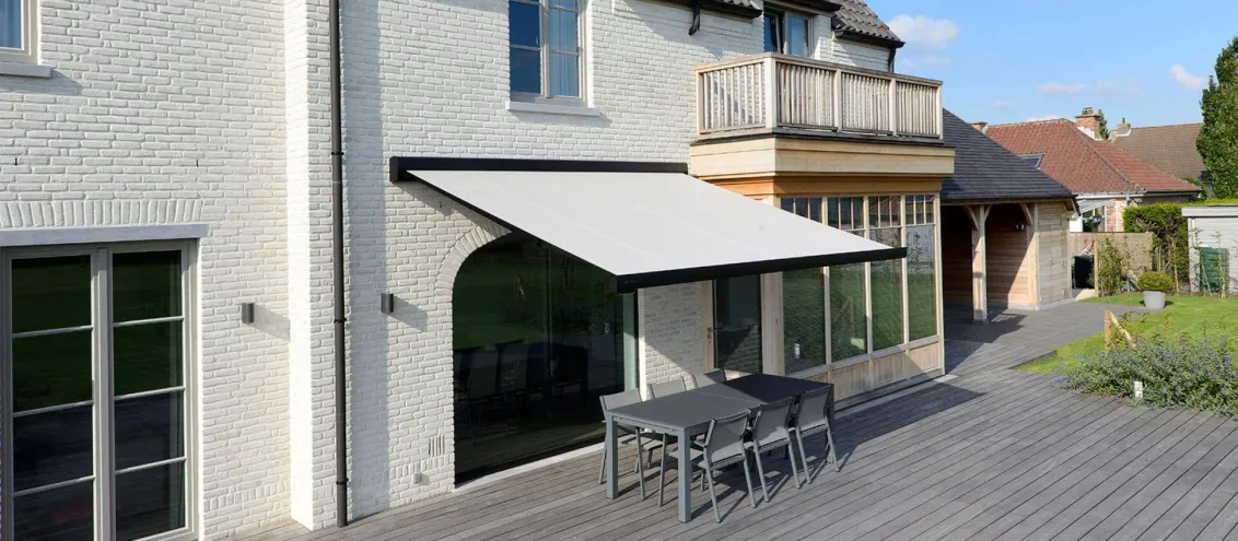 Markise B24 mit grauem Tuch, ausgefahren über einer Holzterrasse mit dunklem Esstisch an einem hellen Klinkerhaus