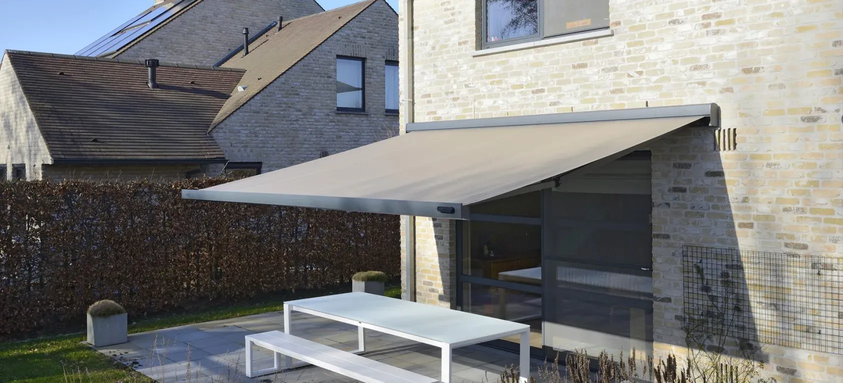 Markise B24 mit grauem Tuch, ausgefahren über einer Terrasse mit hellem Tisch an einem Klinkerhaus, dahinter eine Hecke