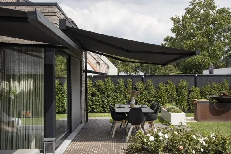 Markise B27 mit anthrazitfarbenem Tuch, ausgefahren über einer Gartenterrasse mit Esstisch und Stühlen, dahinter Hecke und Garten