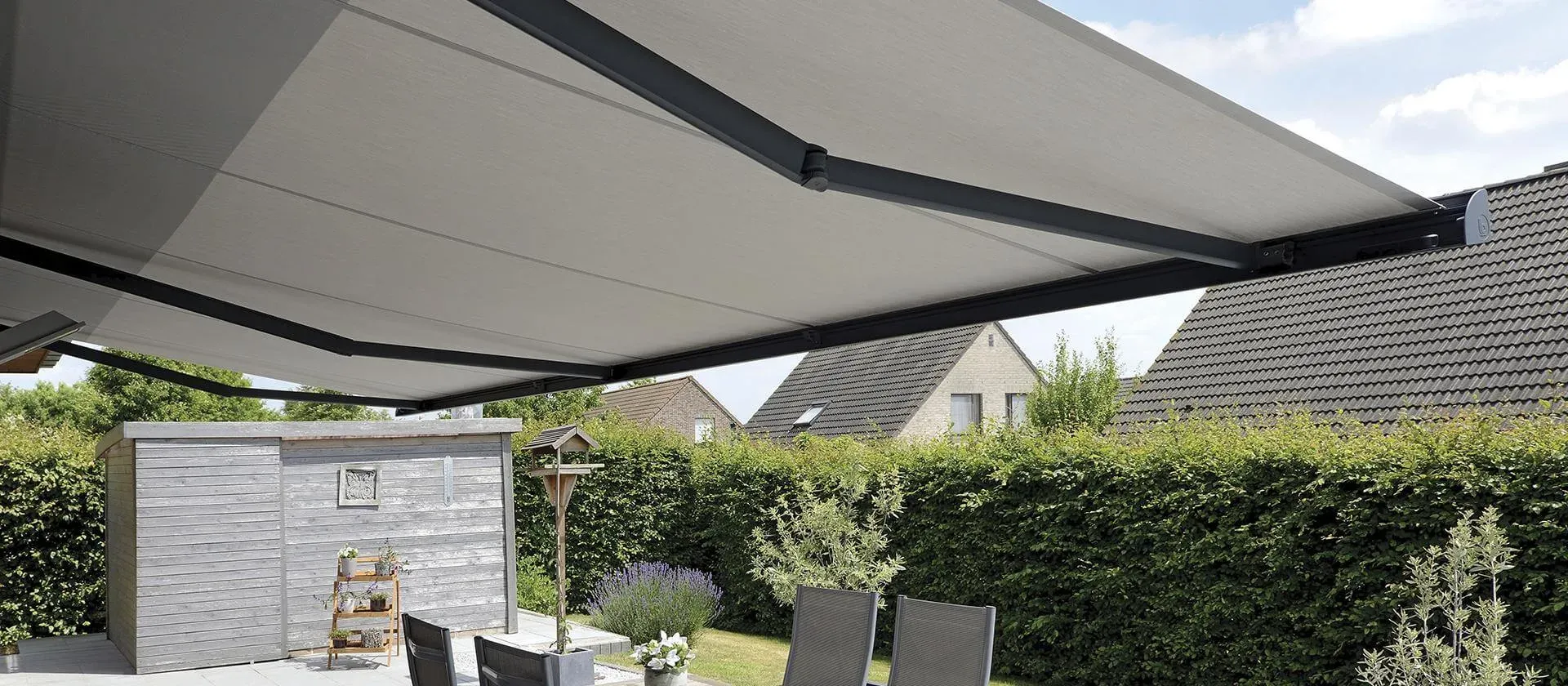 Markise B35 mit grauem Tuch, breit ausgefahren über einer Gartenterrasse mit Stühlen, dahinter eine Hecke
