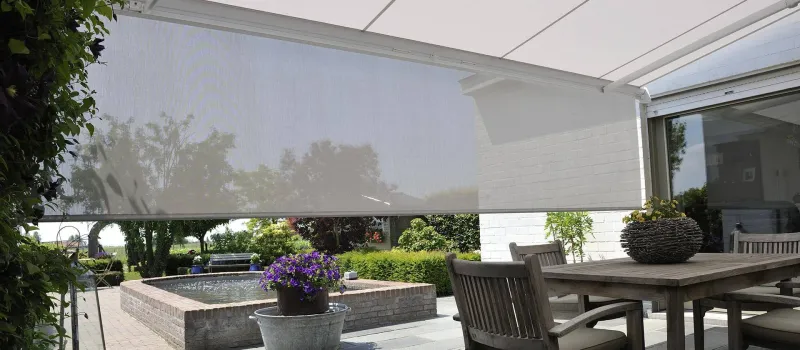 Markise B38 mit heruntergelassenem grauem Senkrechtscreen über einer Terrasse mit Esstisch, dahinter ein Garten mit Wasserbecken
