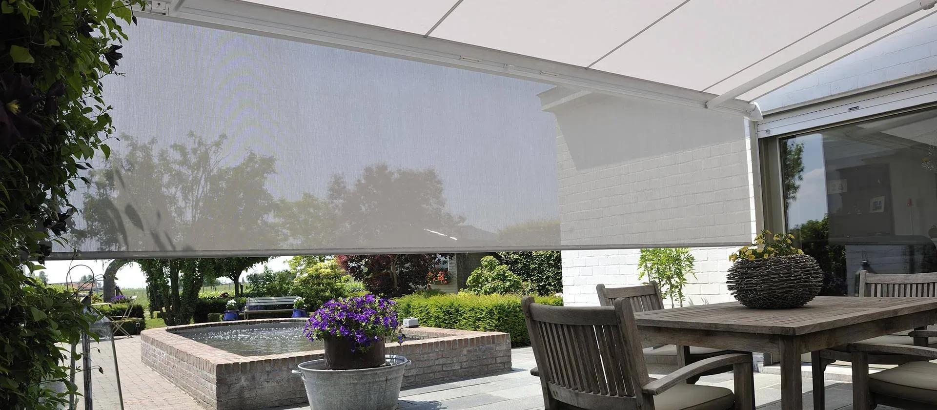 Markise B38 mit heruntergelassenem grauem Senkrechtscreen über einer Terrasse mit Esstisch, dahinter ein Garten mit Wasserbecken