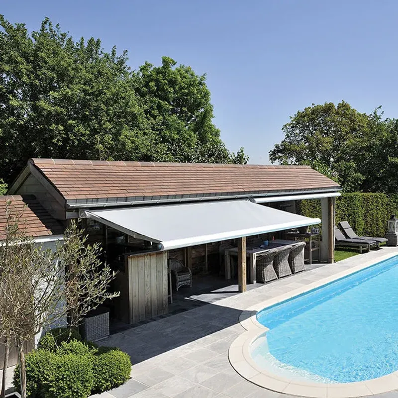 Markise B50 XL mit hellem Tuch, ausgefahren von einem Poolhaus über einem Sitzbereich neben einem Swimmingpool
