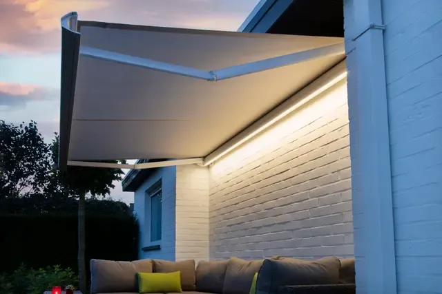 Markise B50 XL mit integrierter LED-Beleuchtung, ausgefahren an einer weissen Klinkerwand über einem Loungebereich in der Abenddämmerung