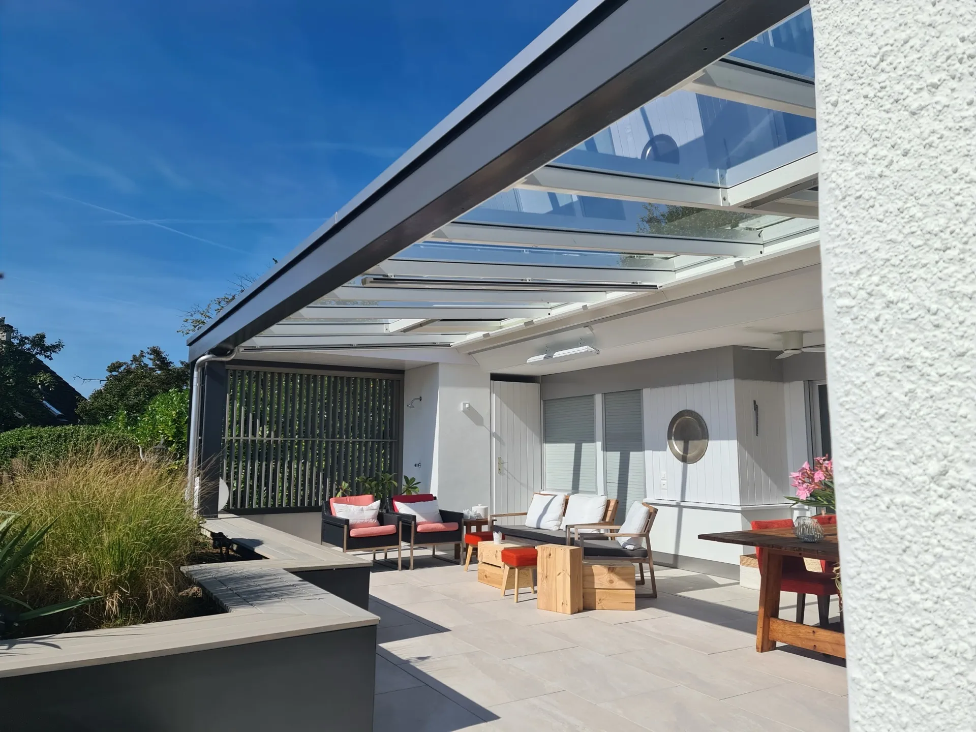 Pergola mit Glasdach über einer Terrasse, dahinter Garten und blauer Himmel